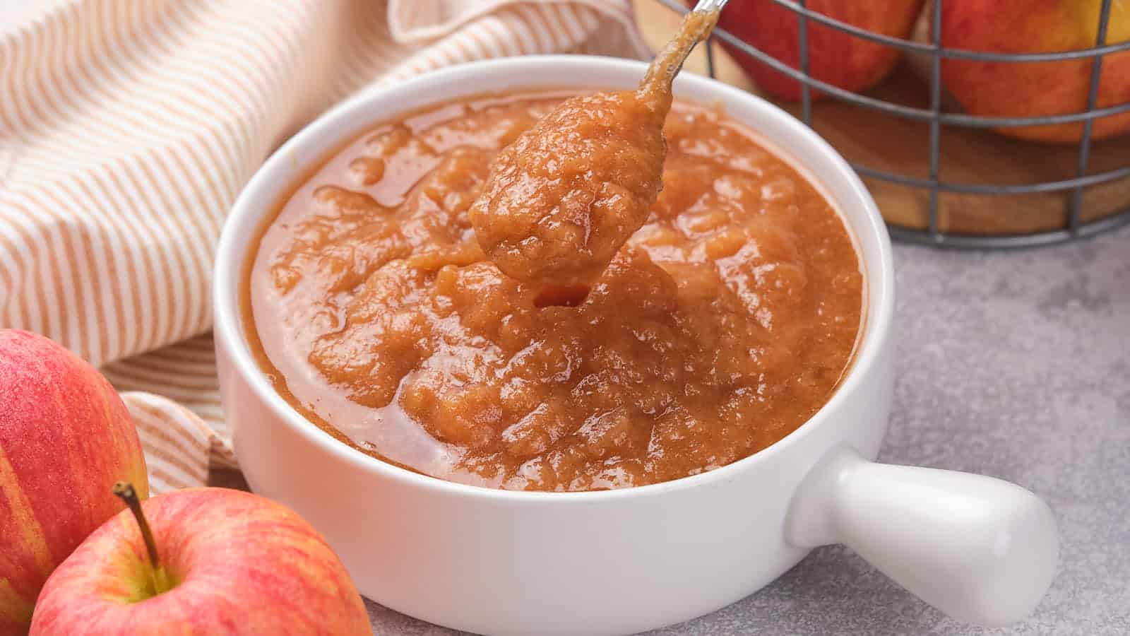 A bowl of applesauce with a spoon, surrounded by apples and a striped cloth on a gray surface.