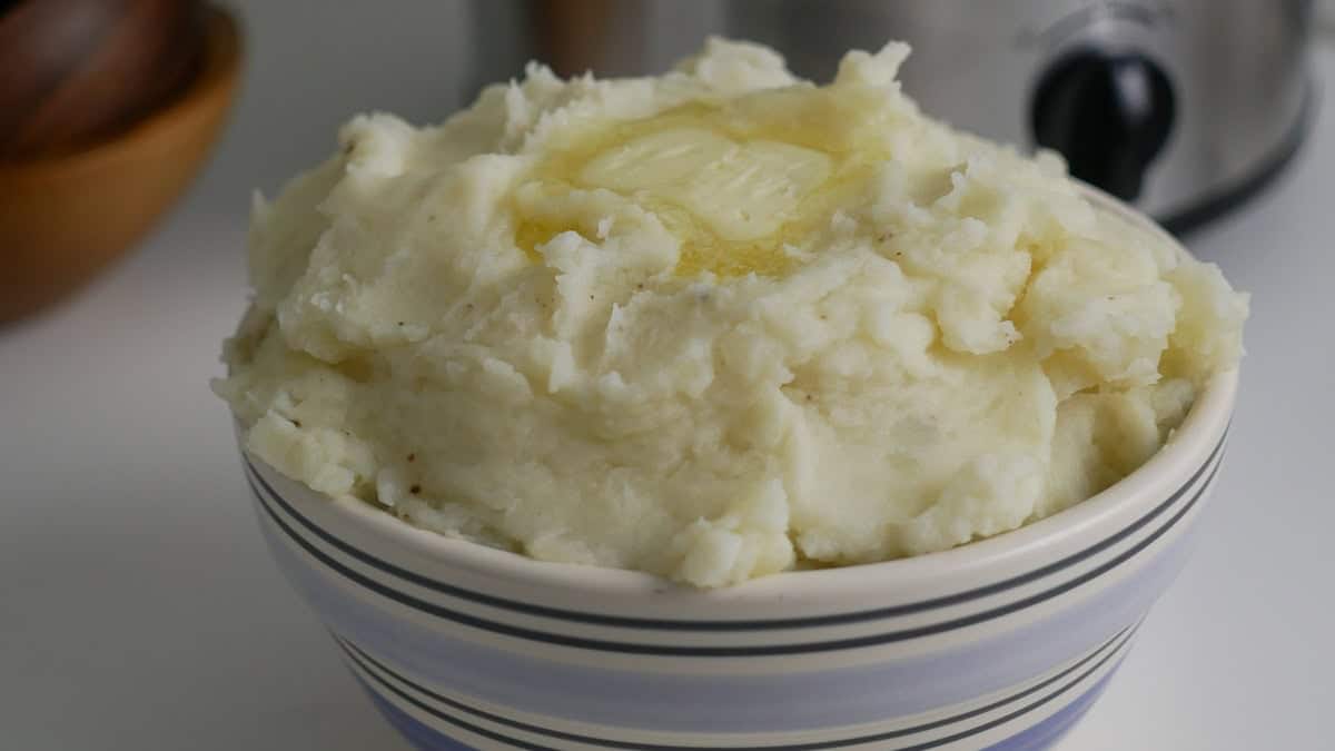 A bowl of mashed potatoes topped with a pat of butter is in front of a slow cooker and some wooden bowls.