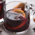 Two clear glass mugs of mulled wine with orange slices, star anise, and cinnamon sticks.