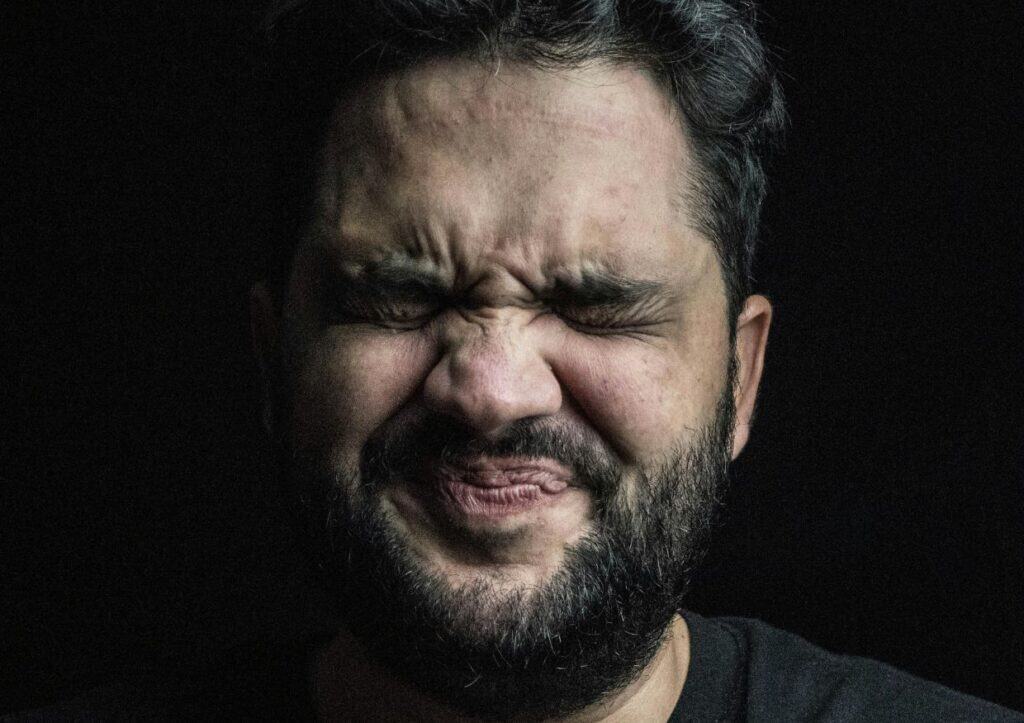 A person with a beard and closed eyes makes a scrunched facial expression against a dark background.