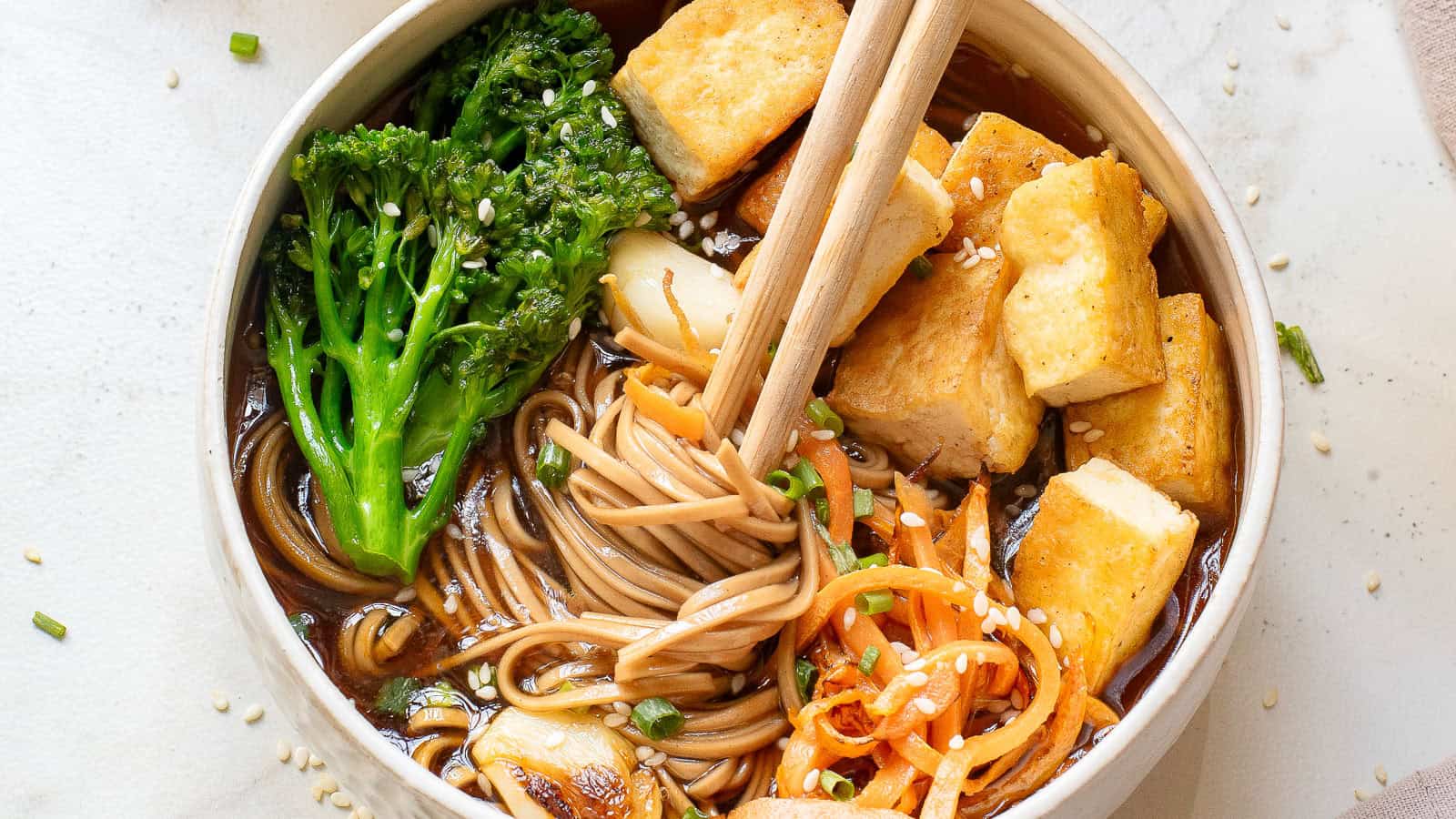 A bowl of soba noodles in broth, topped with fried tofu, broccolini, carrot spirals, and sliced mushrooms. Chopsticks rest on the bowl, and sesame seeds are sprinkled over the dish.