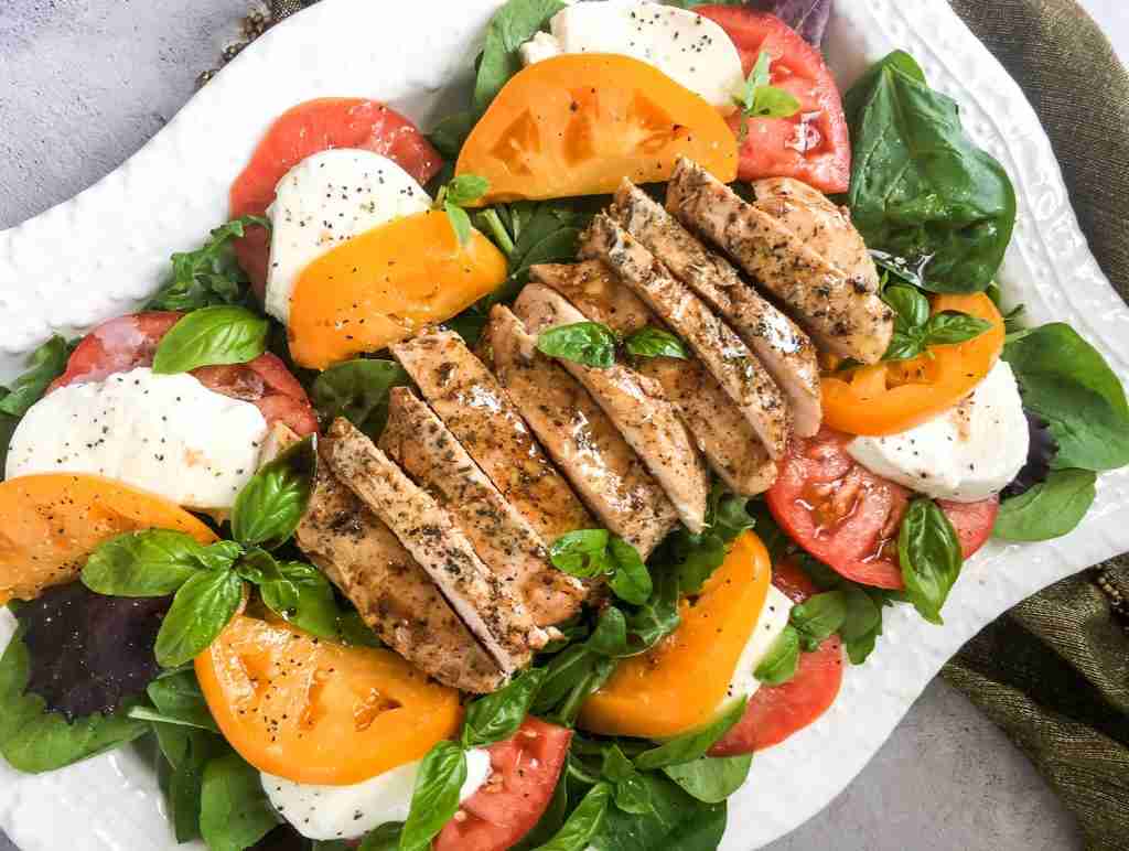 A plate of grilled chicken breast slices on a bed of mixed greens, tomatoes, and mozzarella, garnished with fresh basil leaves.