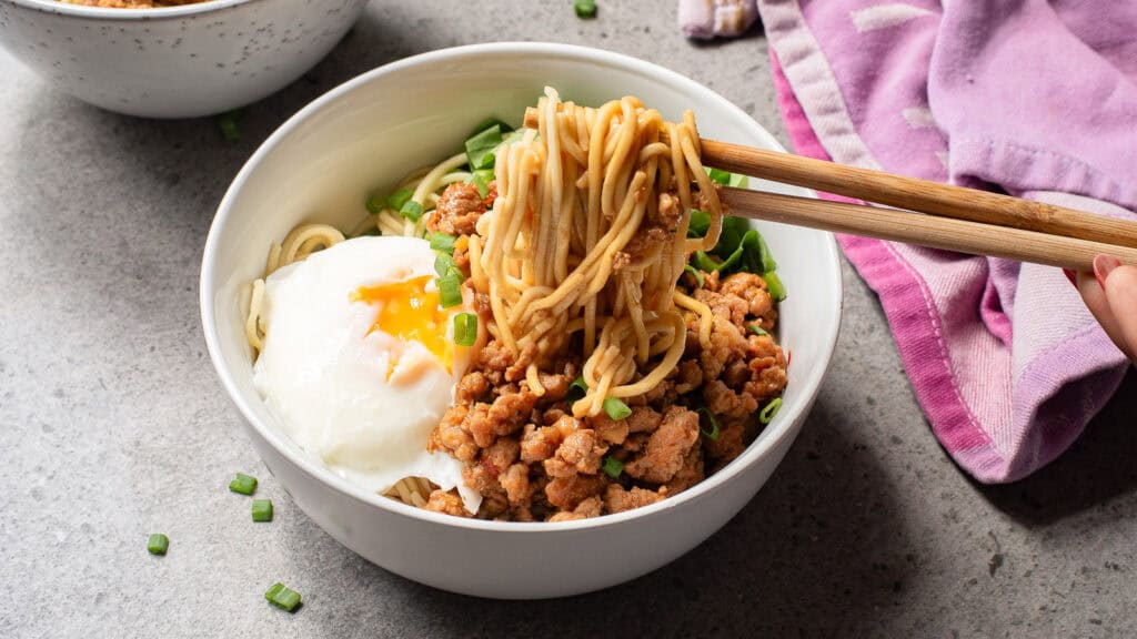 A bowl of noodles with minced meat, a poached egg, and chopped green onions. A hand uses chopsticks to lift the noodles. A purple cloth is partially visible on the side.