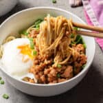 A bowl of noodles with minced meat, a poached egg, and chopped green onions. A hand uses chopsticks to lift the noodles. A purple cloth is partially visible on the side.