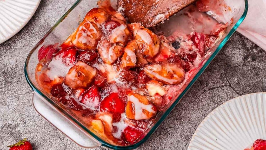 A glass baking dish with partially served strawberry cobbler, showing a mix of strawberries and biscuit topping. Two empty plates are nearby.