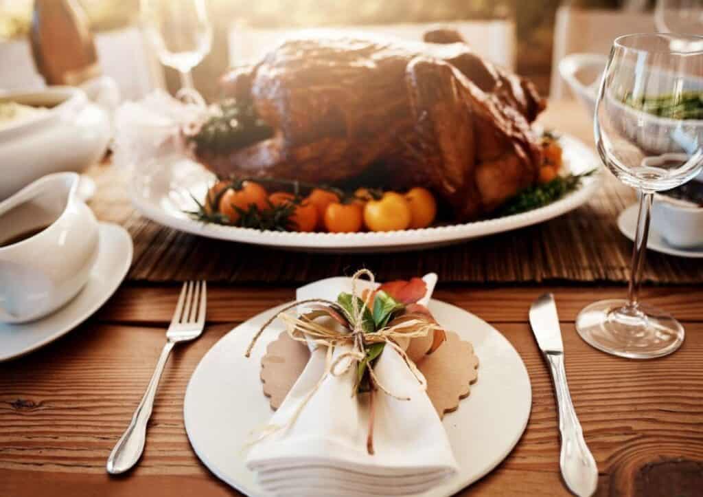 A roasted turkey on a platter with tomatoes and herbs offers the perfect centerpiece for a stress-free Thanksgiving dinner. A dining table is set, complete with a fork, knife, and a white napkin tied with twine, ready to welcome family and friends.