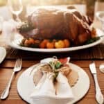 A roasted turkey on a platter with tomatoes and herbs offers the perfect centerpiece for a stress-free Thanksgiving dinner. A dining table is set, complete with a fork, knife, and a white napkin tied with twine, ready to welcome family and friends.