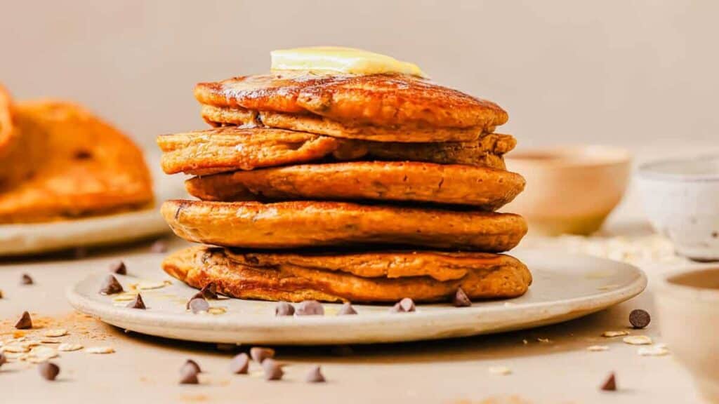 A stack of pancakes topped with a slice of butter sits on a plate. Chocolate chips and oats are scattered around the plate.