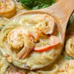 A wooden spoon scoops up a portion of creamy shrimp pasta with red bell peppers and fresh basil, garnished with a lime wedge.