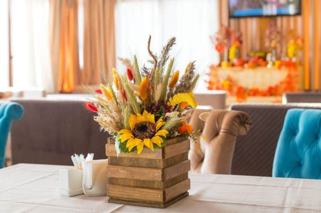 A wooden box adorned with a sunflower and various dried flowers sits on a table with blue chairs, surrounded by fall-themed decor in warm Thanksgiving colors.