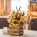 A wooden box adorned with a sunflower and various dried flowers sits on a table with blue chairs, surrounded by fall-themed decor in warm Thanksgiving colors.