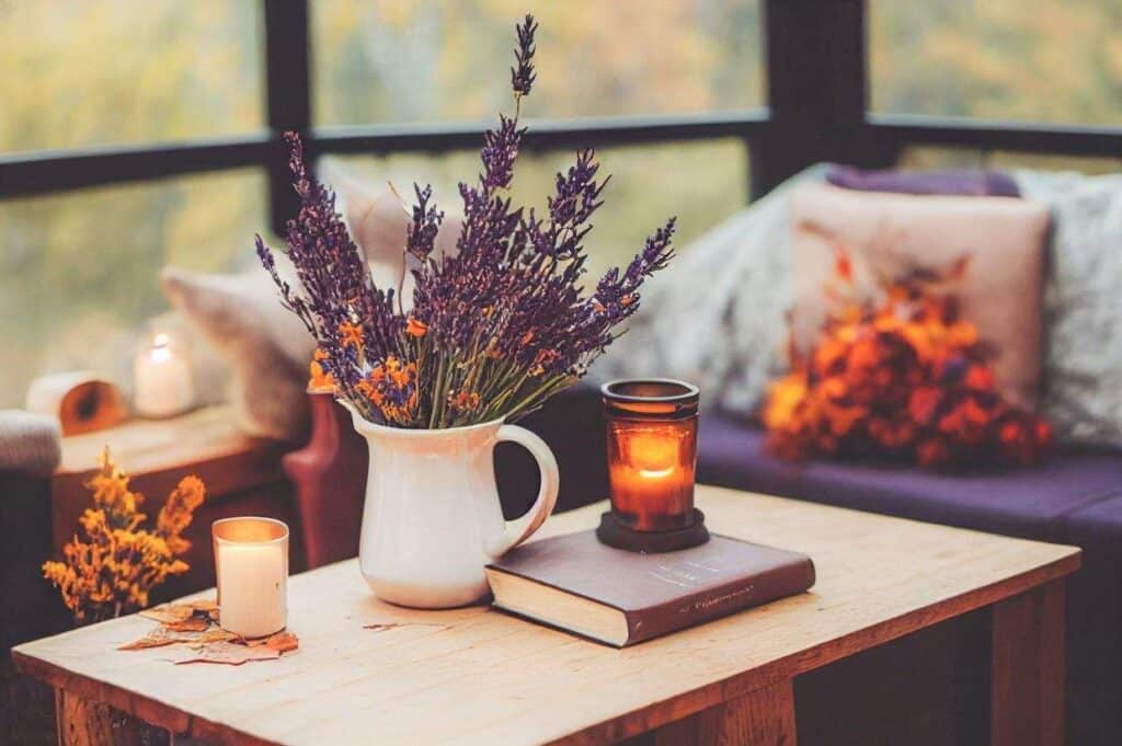 A cozy living room scene showcases a wooden table adorned with a white pitcher of lavender, a candle resting on a book, and autumn-themed decor in thanksgiving colors, all enveloped in a softly lit ambiance.