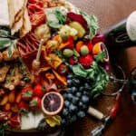 A colorful cheese board with meats, cheeses, fruits, and vegetables is placed next to a bottle and glass of red wine on a wooden table.