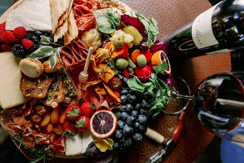 A colorful cheese board with meats, cheeses, fruits, and vegetables is placed next to a bottle and glass of red wine on a wooden table.