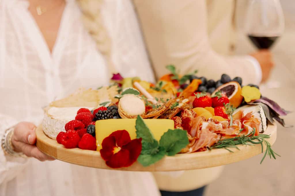 A person holding a wooden platter with assorted cheeses, meats, fruits, berries, and a wine glass in the background.