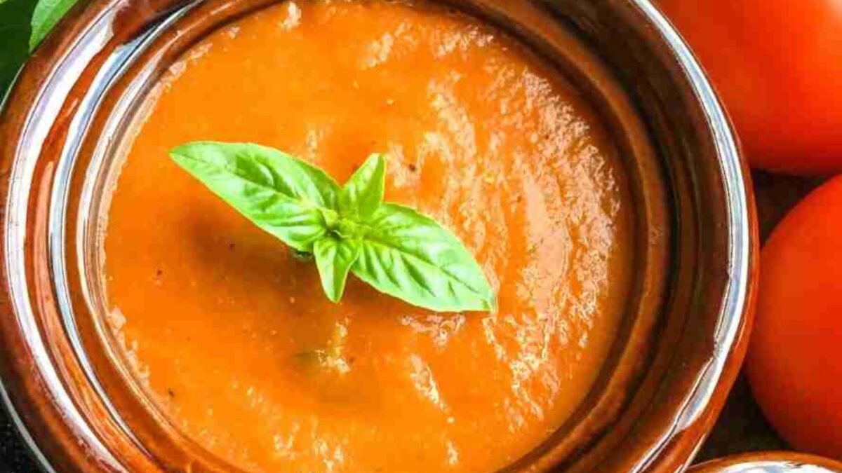 A bowl of tomato soup garnished with a basil leaf, next to fresh tomatoes.