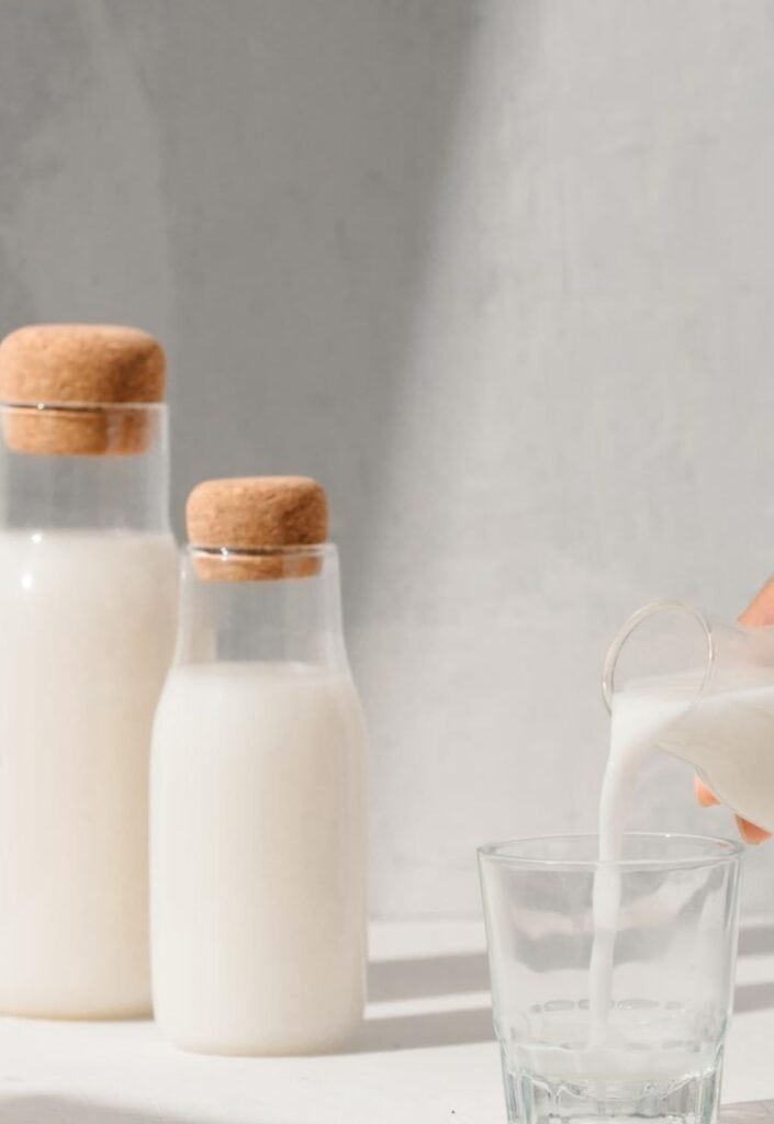 Two bottles of milk with cork stoppers and a hand pouring milk into a glass on a light background.