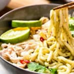 Bowl of noodles with shredded chicken, lime slices, cilantro, and slices of chili, being picked up with chopsticks.