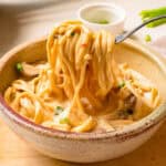 A fork lifts creamy noodles with vegetables from a ceramic bowl on a wooden board.