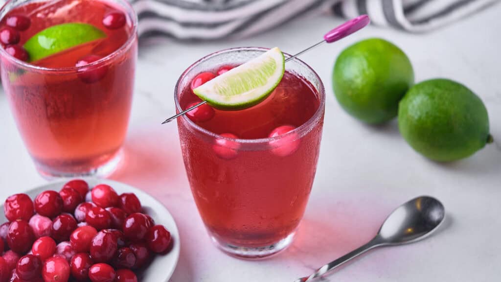 Two glasses of red cranberry cocktail garnished with lime wedges.