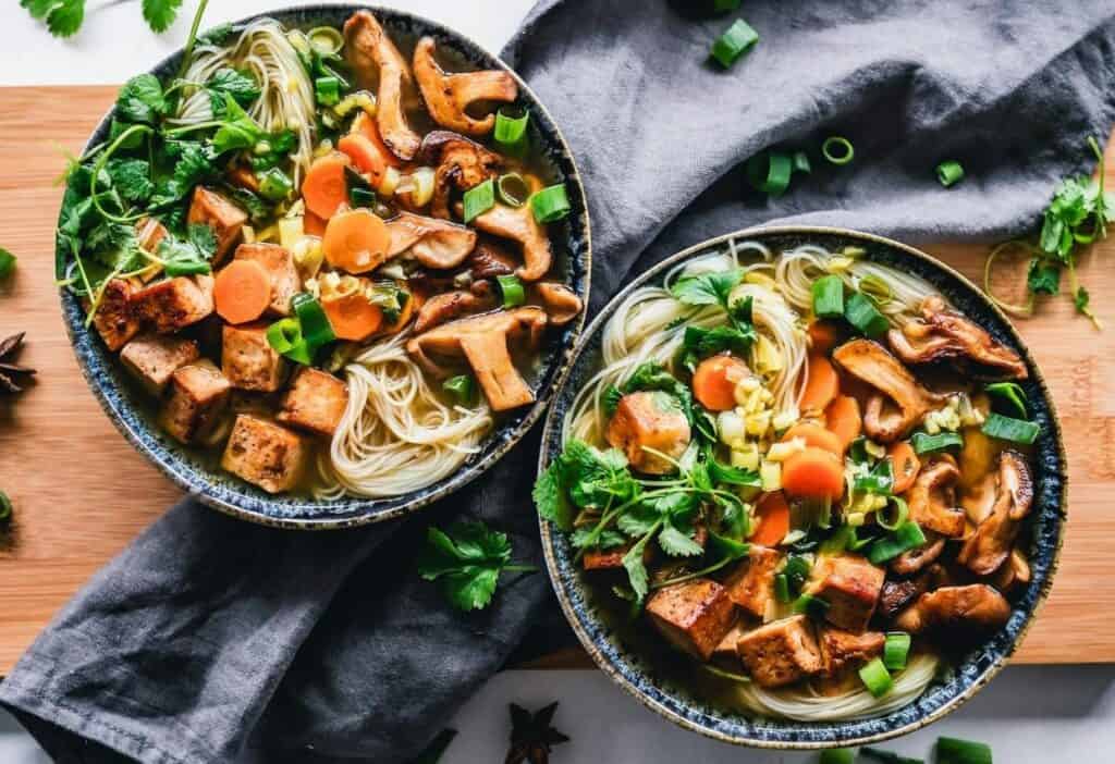 Overhead view of two bowls of noodle soup with vegetables, mushrooms, tofu, green onions, and cilantro on a wooden surface with a grey cloth.