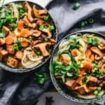 Overhead view of two bowls of noodle soup with vegetables, mushrooms, tofu, green onions, and cilantro on a wooden surface with a grey cloth.