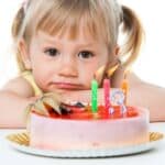 A young child with blonde hair looks at a birthday cake with lit candles displaying the number three.