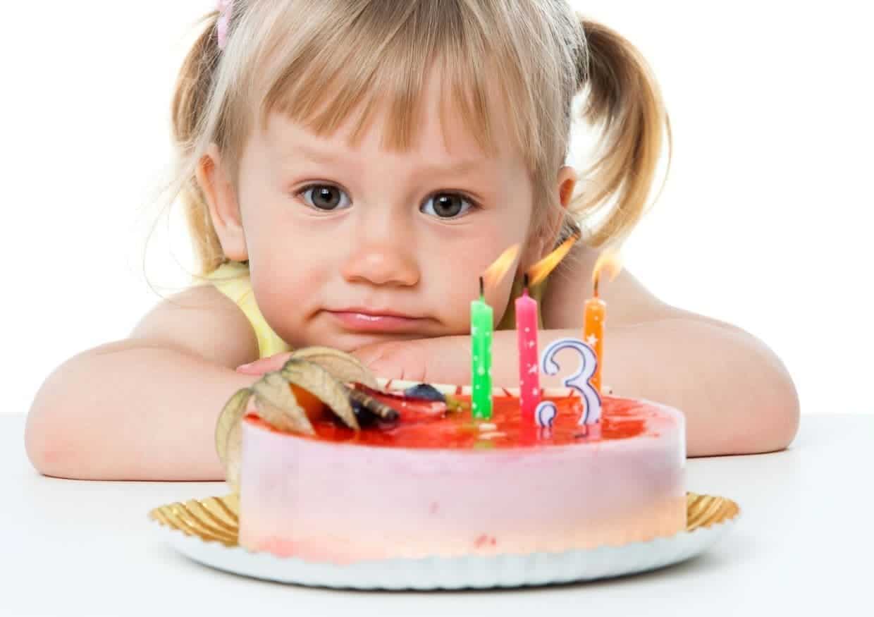 A young child with blonde hair looks at a birthday cake with lit candles displaying the number three.