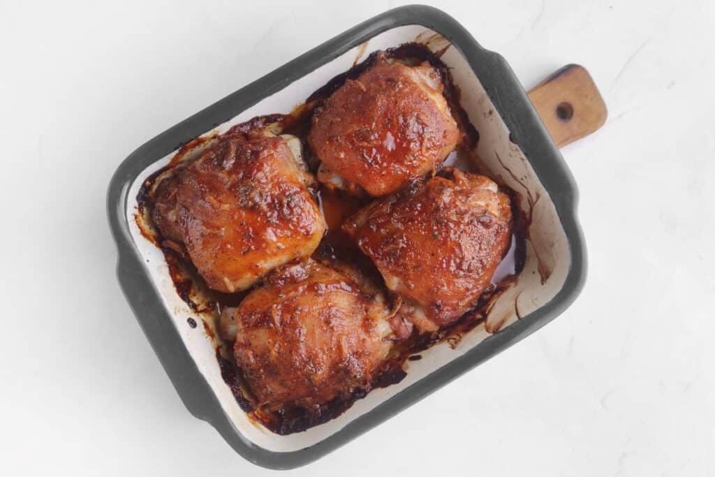 Baked chicken thighs with a caramelized glaze in a rectangular roasting pan on a white surface.