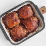 Baked chicken thighs with a caramelized glaze in a rectangular roasting pan on a white surface.