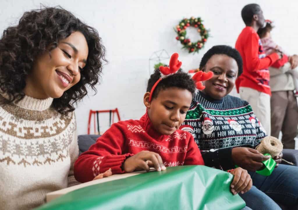 A child wearing reindeer antlers unwraps a gift with two adults beside them. A family is gathered in the background, dressed in festive sweaters and attire.