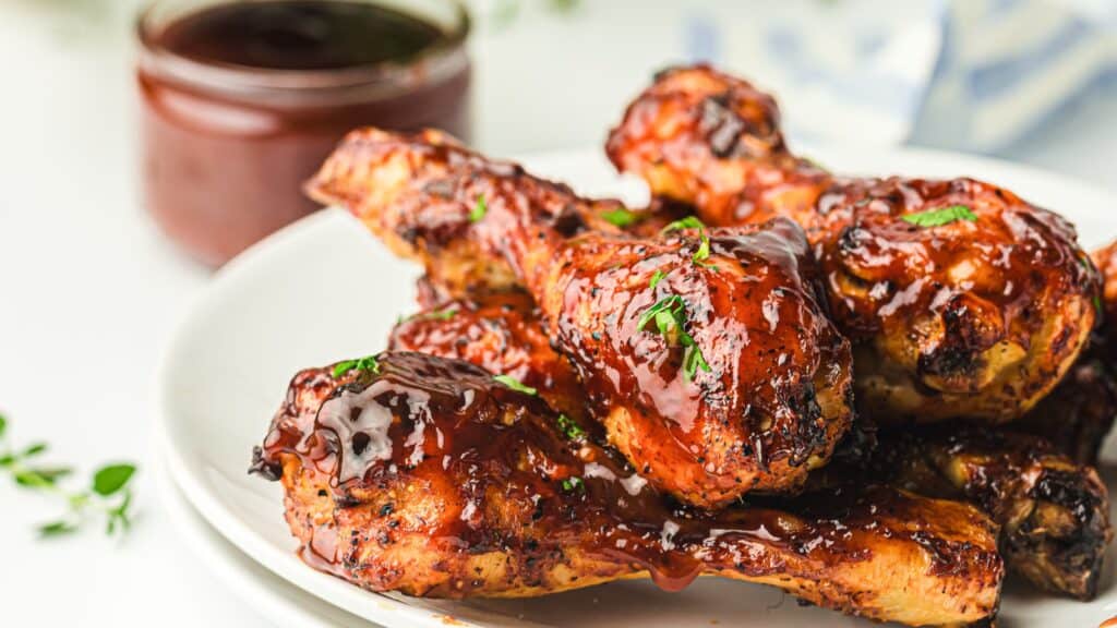 A plate of grilled chicken drumsticks covered in glossy barbecue sauce, garnished with herbs. A jar of sauce is visible in the background.