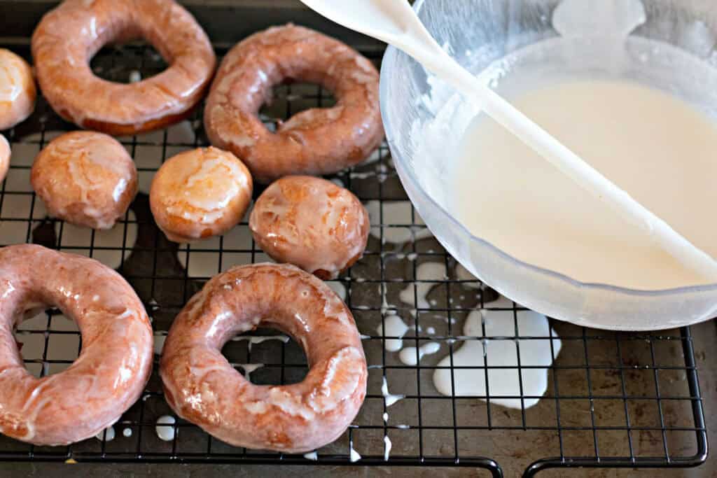 Glazed donuts and donut holes on a cooling rack beside a bowl of white icing with a spatula.