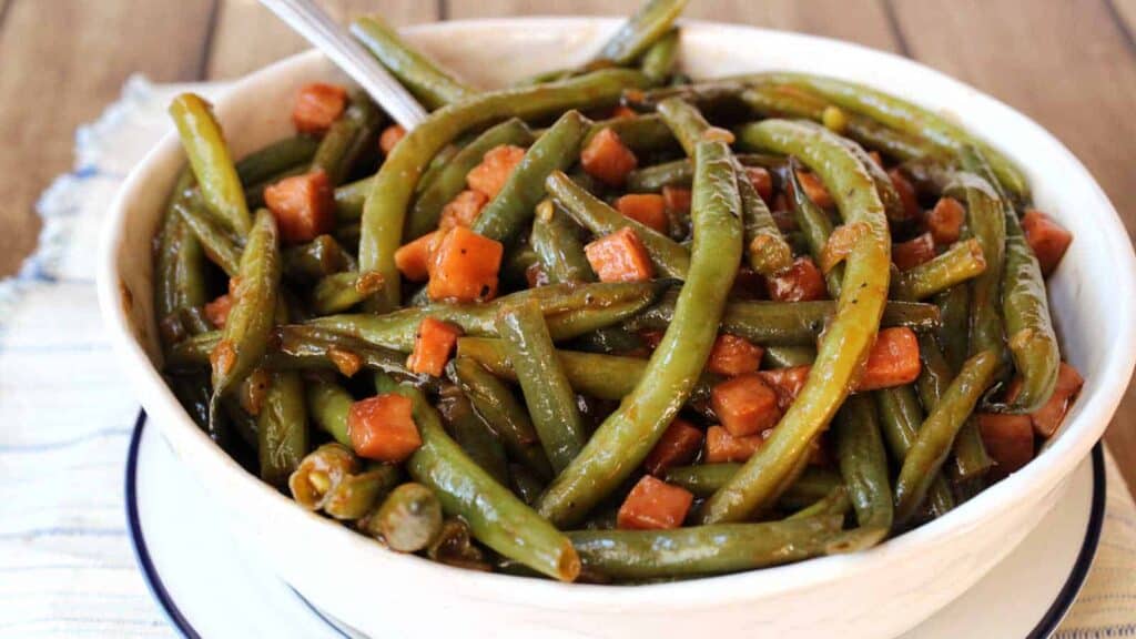 A bowl of cooked green beans mixed with diced ham and a sauce. A spoon rests in the bowl.