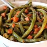 A bowl of cooked green beans mixed with diced ham and a sauce. A spoon rests in the bowl.