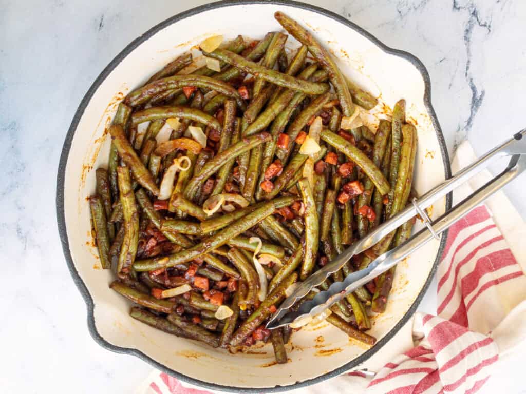 A skillet with cooked green beans, diced onions, and small sausage pieces. Tongs rest on the dish's edge. A striped cloth partially visible underneath.