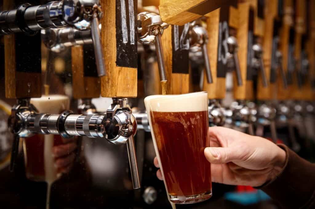 A person pours a glass of beer from a row of taps, with wooden handles, into a glass with a foamy head.