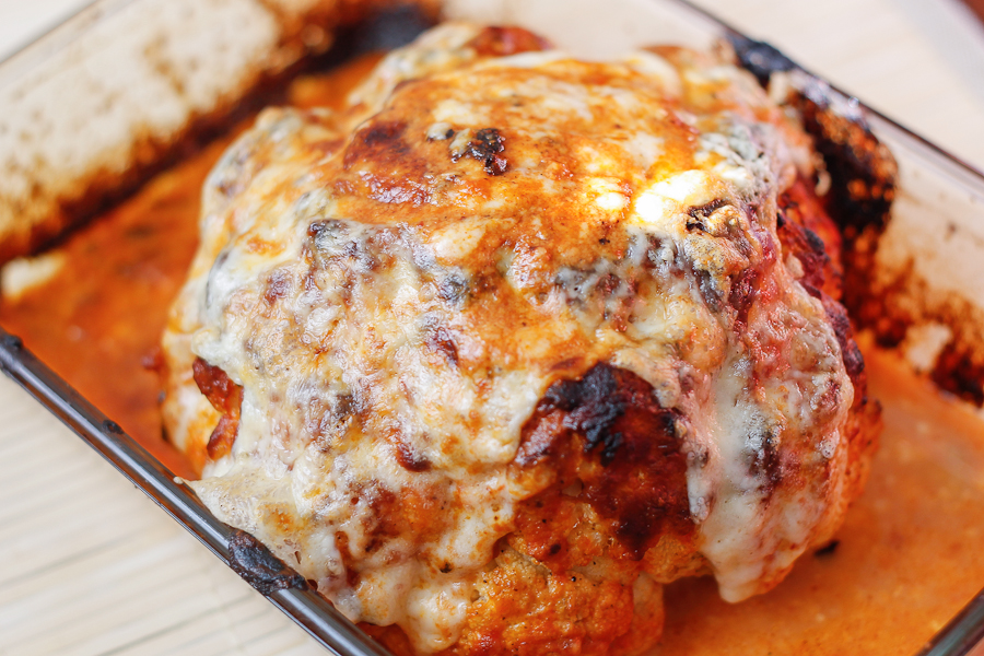 A whole roasted cauliflower covered with melted cheese and sauce, in a baking dish.