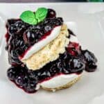 A dessert with two layers of pastry, cream, and blueberry sauce, topped with fresh mint, on a white square plate.