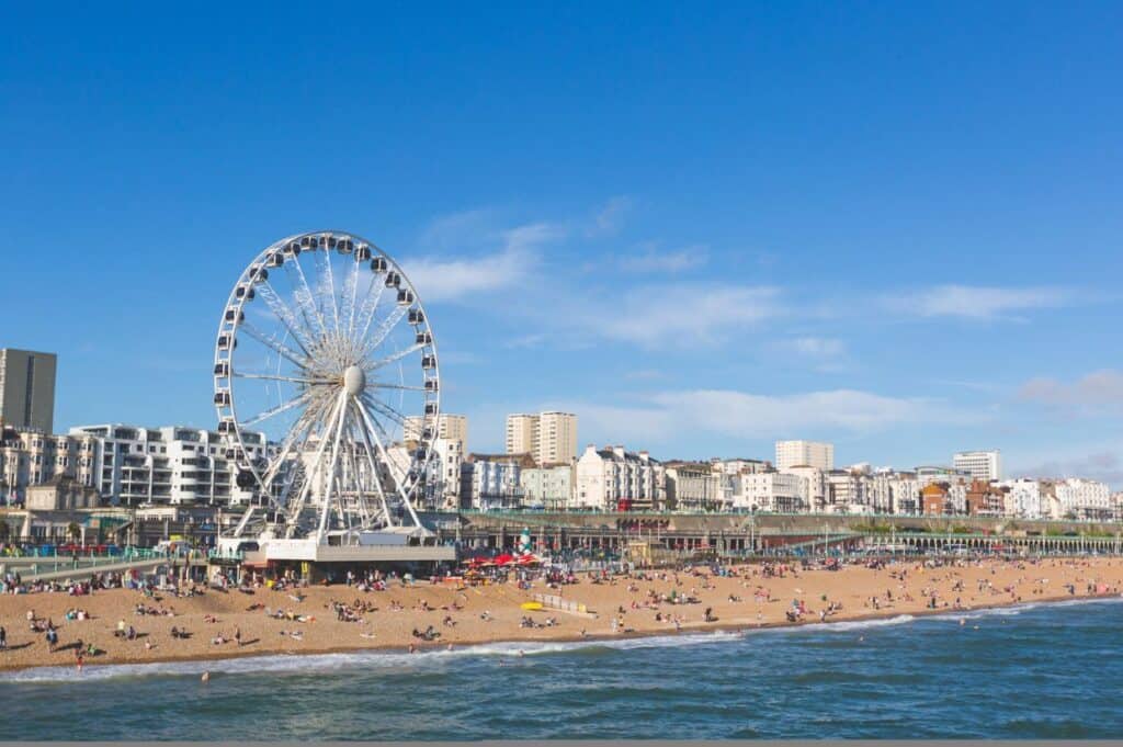 A large Ferris wheel towers over a busy beach where people unwind on the sand. This scenic spot, perfect for day trips from London, is complemented by a cityscape and a clear blue sky in the background.