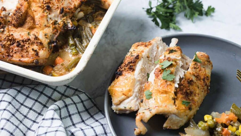 Grilled chicken breasts on a gray plate, garnished with herbs, with a baking dish of roasted vegetables in the background. A checkered cloth is in the foreground.