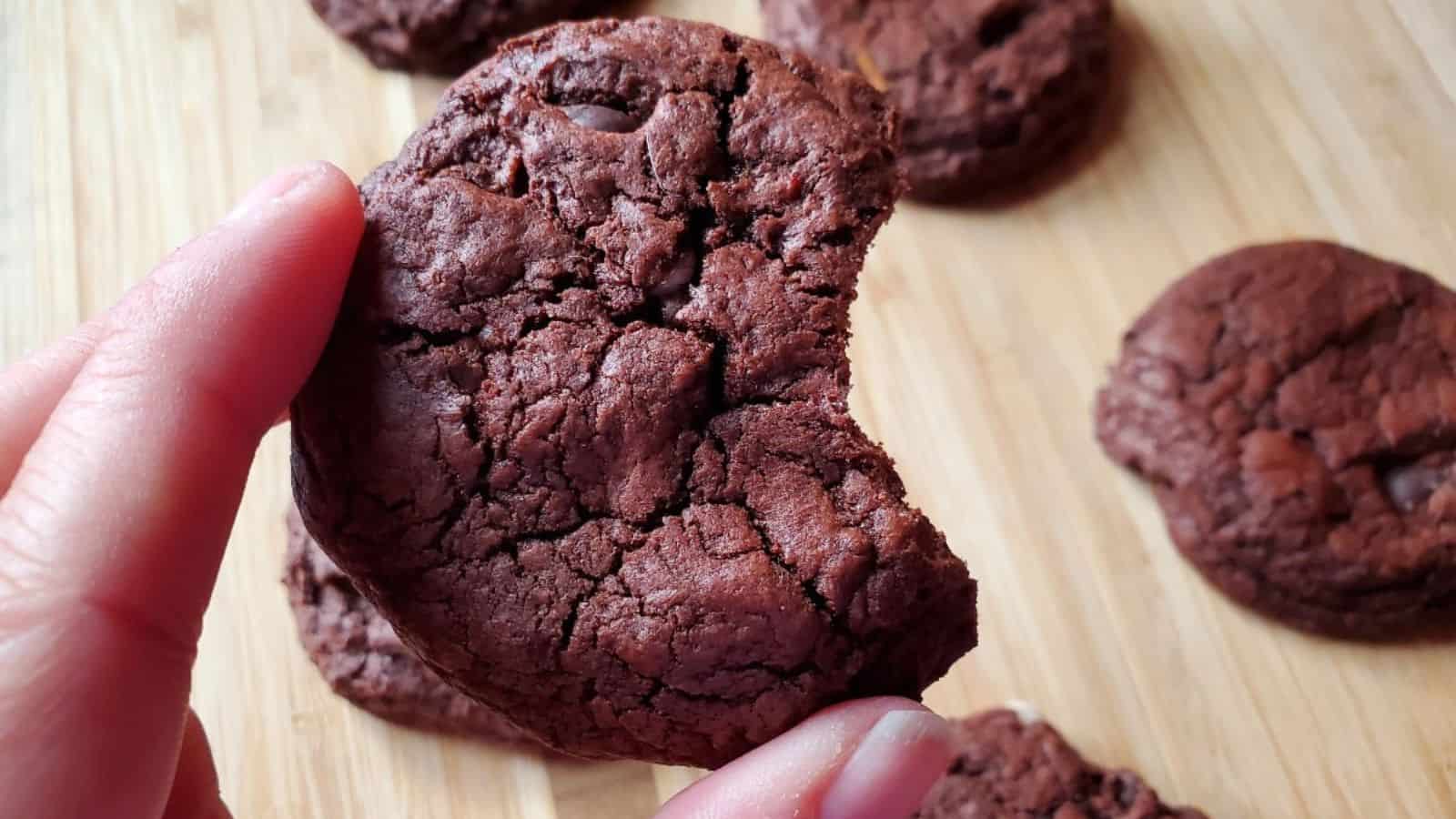 Close-up of a hand holding a bitten chocolate cookie with more cookies in the background on a wooden surface.