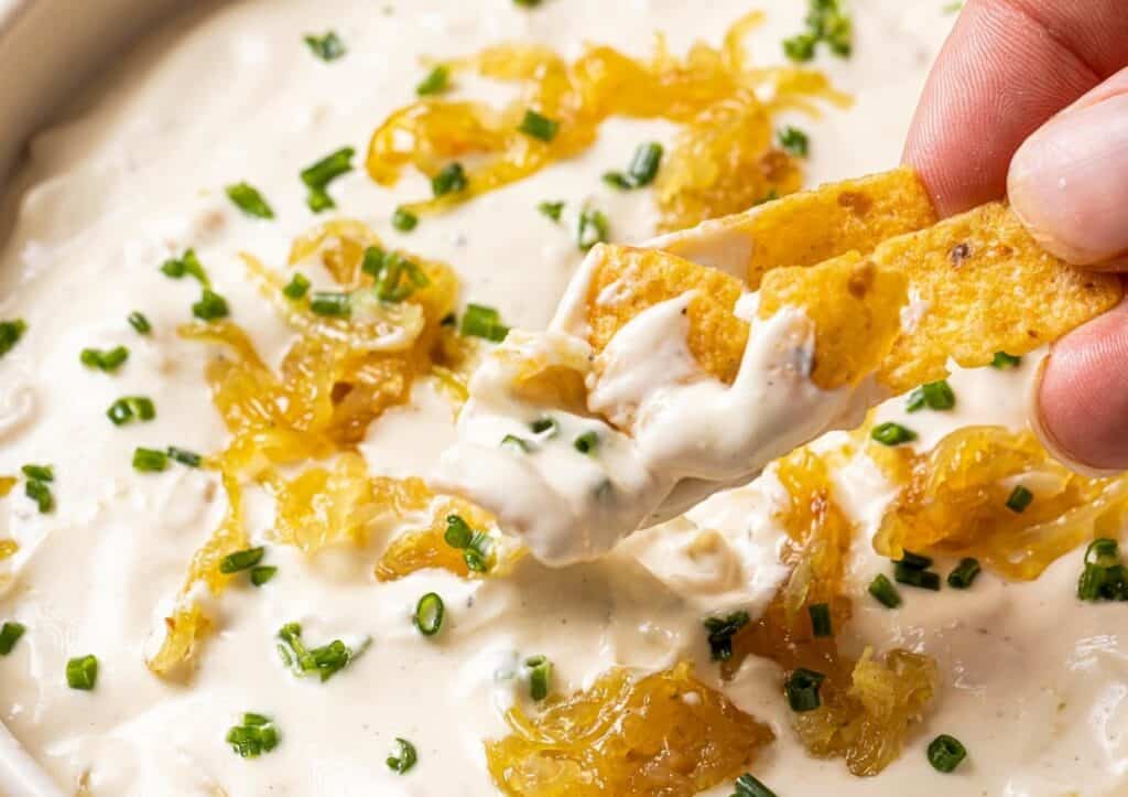 A tortilla chip dipped in creamy white onion dip, garnished with caramelized onions and chopped chives.