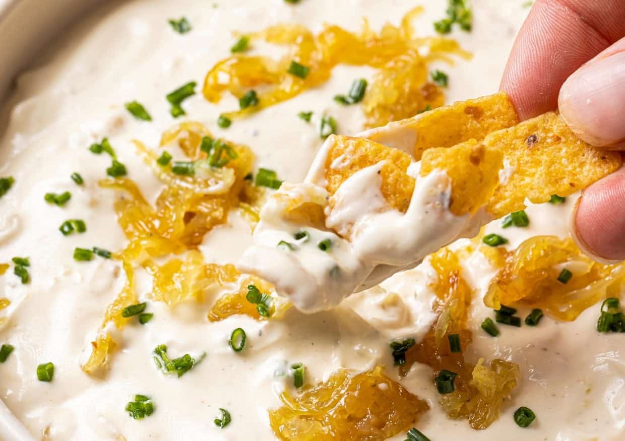 A hand dips a tortilla chip into creamy onion dip topped with caramelized onions and chopped chives. The dip is white and thick, with visible onion pieces and green chive garnishes spread across the surface.