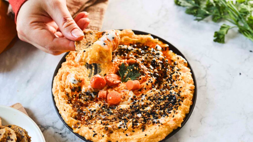 A hand dips a cracker into a bowl of hummus topped with sliced carrots, sesame seeds, and drizzled sauce. Parsley is visible in the background on a marble surface.