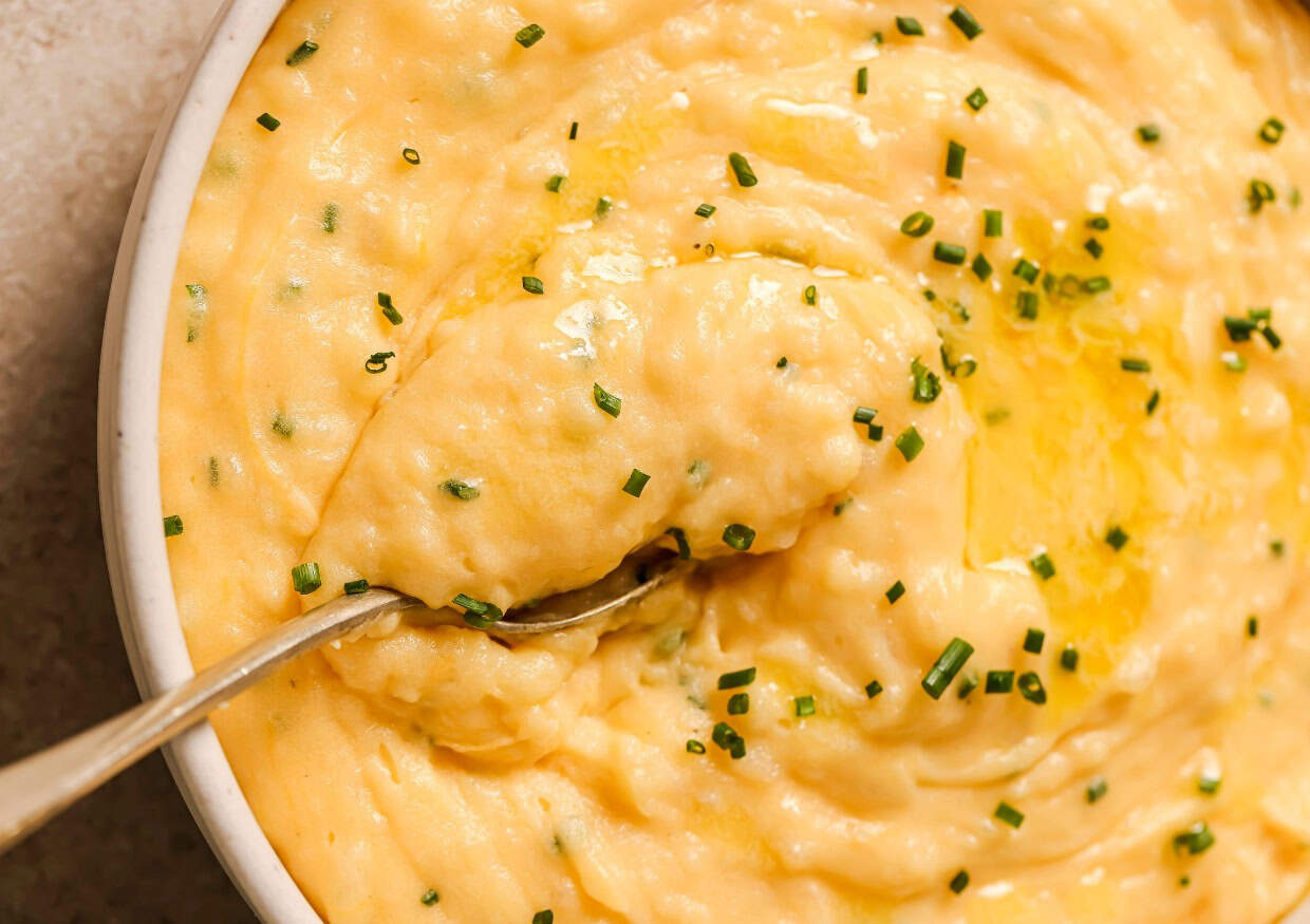 A close-up of creamy mashed potatoes topped with melted butter and sprinkled with chopped chives. A spoon is partially submerged in the mashed potatoes, showcasing their smooth and fluffy texture.