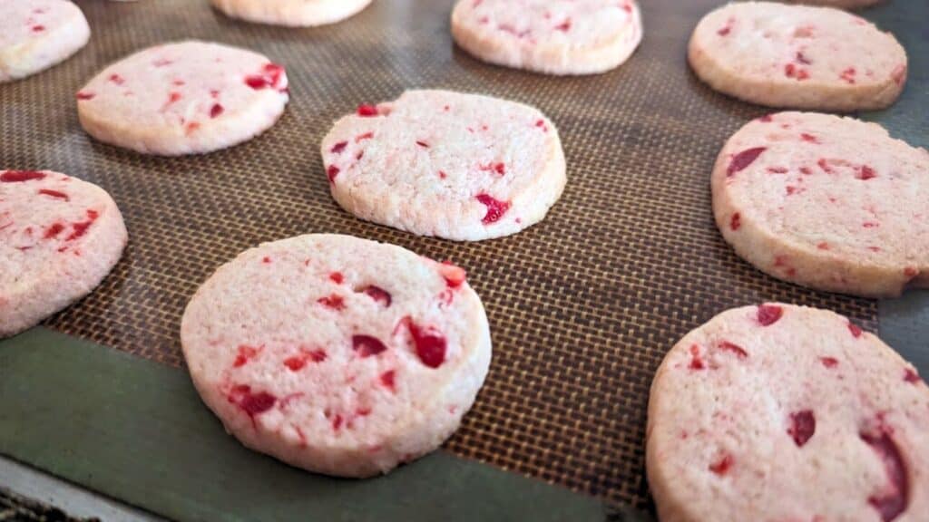 Round cookies with red berry pieces are lined up on a baking mat.