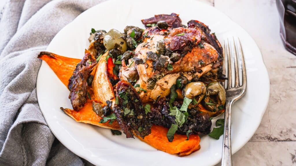 A plate with roasted chicken, sweet potato wedges, olives, and herbs. A fork rests beside the food.