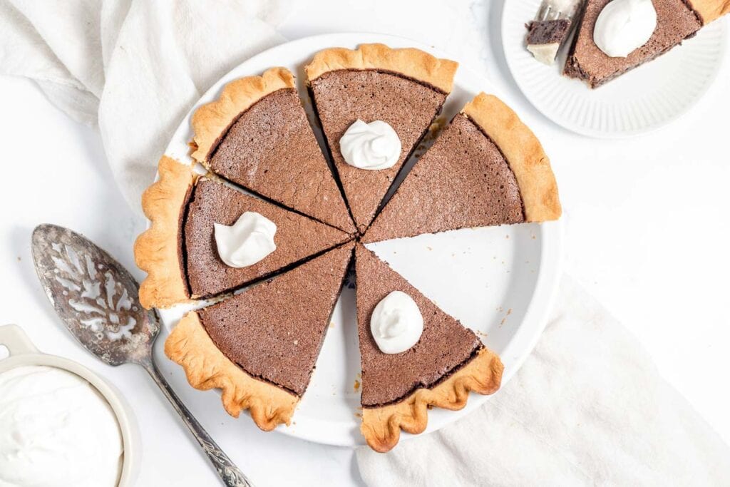 A chocolate pie sliced into five pieces, each topped with a dollop of whipped cream, on a white plate. A cake server, a bowl of whipped cream, and a small dessert plate are nearby.