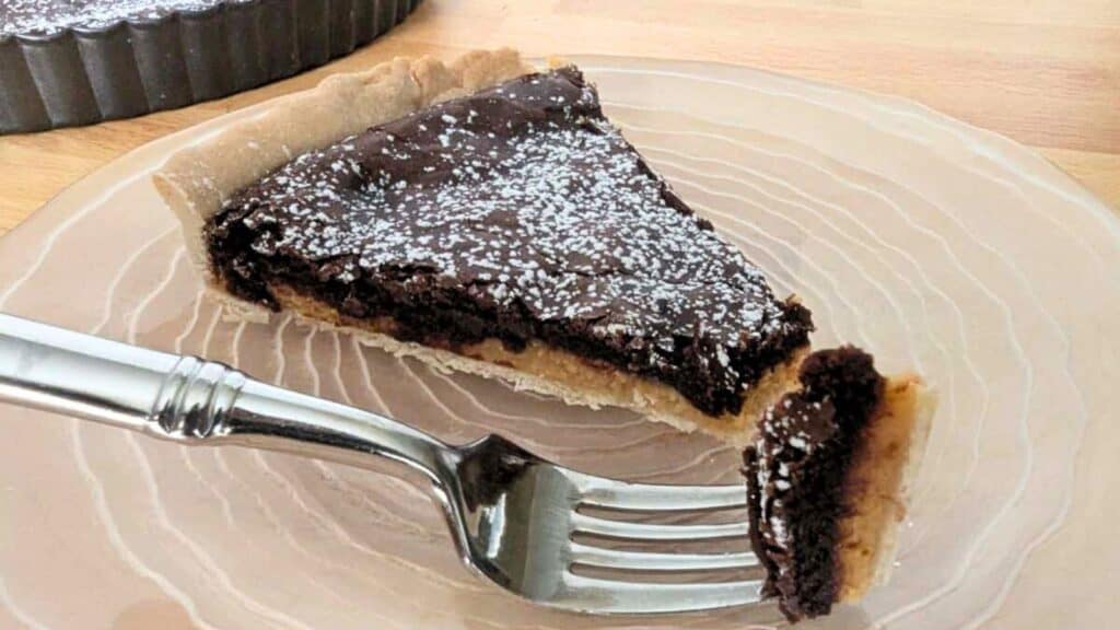 A slice of chocolate tart on a clear plate with a fork. The tart is topped with powdered sugar.